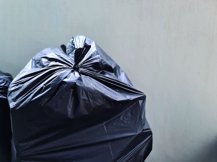 A small, light blue pickup truck is parked on the side of a street, heavily loaded with various types of rubbish and discarded items. The cargo area is filled with a mix of cardboard boxes, black plas
