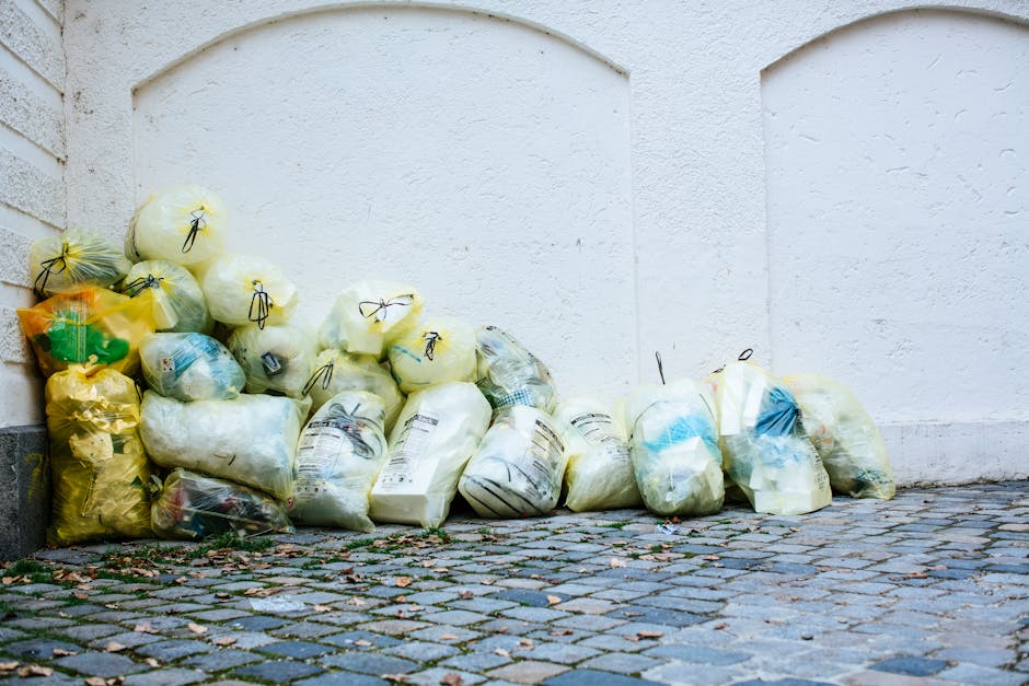 A collection of multiple clear and yellow plastic rubbish bags filled with waste is piled against a white brick wall, situated on a paved cobbled street surface. The bags vary in size, some appearing