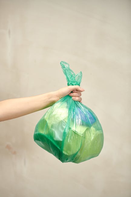A collection of multiple clear and yellow plastic rubbish bags filled with waste is piled against a white brick wall, situated on a paved cobbled street surface. The bags vary in size, some appearing